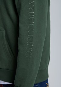 Sudadera con capucha de color verde oscuro con una textura suave, que presenta un logotipo en relieve a lo largo de la manga izquierda. Incluye un bolsillo frontal y puños acanalados.
