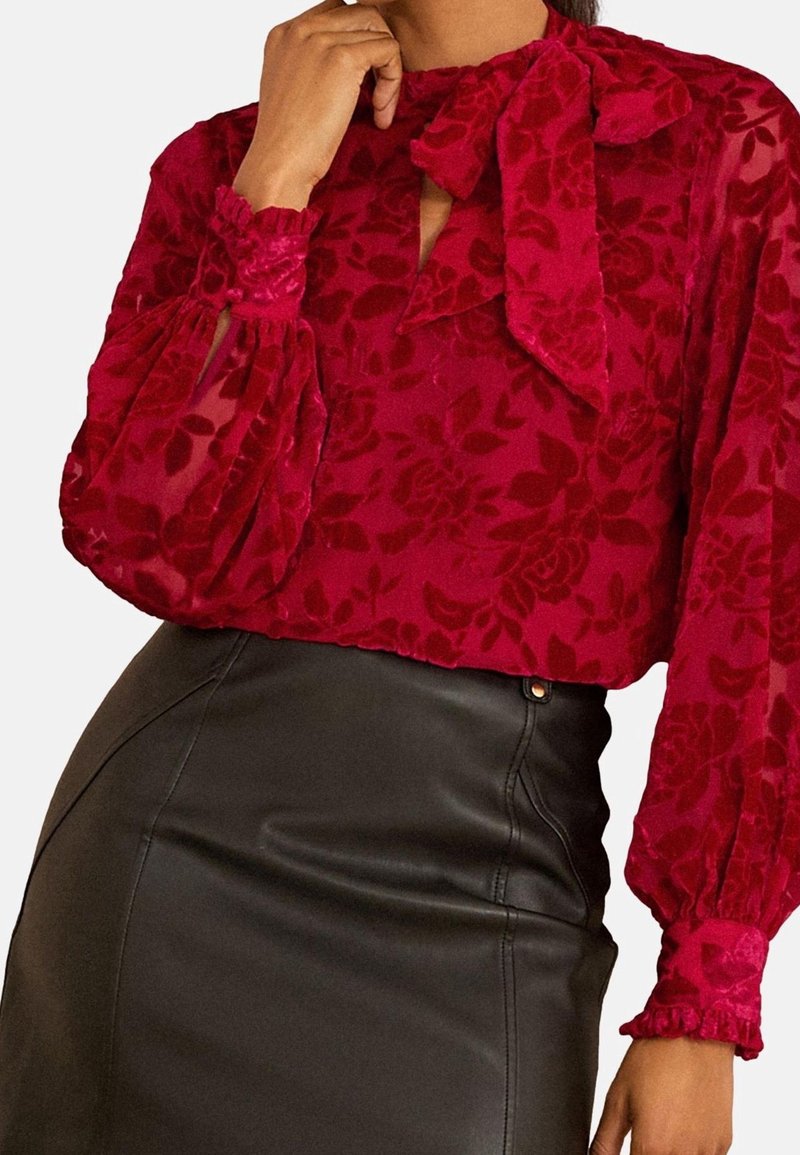 Femme portant un chemisier en velours rouge à fleurs avec de longues manches bouffantes et un nœud au col, associé à une jupe en cuir noire taille haute.