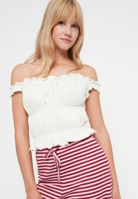 Blusa branca de ombros à mostra com bordas franzidas, cintura elástica; combinada com calções às riscas vermelhas e brancas com cordão de ajustamento.