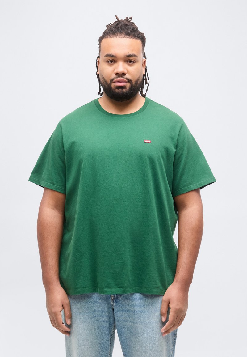 Άνδρας με πλεξούδες στα μαλλιά και γενειάδα, φορώντας πράσινο t-shirt Levi's και ανοιχτόχρωμο μπλε τζιν, στέκεται μπροστά σε απλό λευκό φόντο.