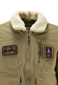 Giacca bomber verde con colletto in finta pelliccia, cerniere e toppa in stile militare raffigurante un'aquila e un emblema tricolore.