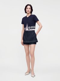 Femme portant un cardigan court bleu marine à manches courtes avec une bordure florale blanche, un short noir taille haute et des talons beiges, debout avec une main dans la poche.