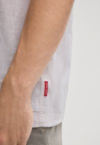 Camicia bianca di lino con una texture sottile, dotata di un'etichetta rossa sul lato. Un braccio è visibile, poggiato su una superficie chiara.