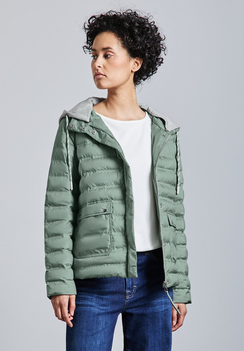 Grön pufferjacka med en grå huva, som har dragkedja fram, sidfickor och ett kviltat mönster. Avslappnad passform, bärs över en vit topp.