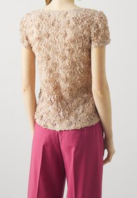 Beige květinový top s aplikacemi a měkkou texturou, s krátkými rukávy, spárovaný s růžovými kalhotami. Top má složité vrstvené designy růží.