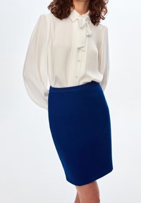 Vit blus med knytband och knapplist, kombinerad med en figurnära blå kjol. Blusen har puffärmar; kjolen har en slät textur.