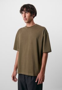 Oliven grøn bomulds T-shirt med rund hals, korte ærmer, afslappet pasform og syede kanter, præsenteret på en model med lys hudtone.