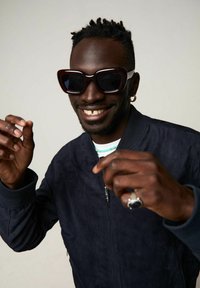 Sunglasses med stora mörka linser, tjocka vinröda bågar med vita accenter. Modellen bär en mörk bomberjacka och en silverring, med armarna lyfta.