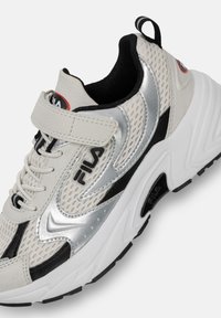 Baskets de sport blanches avec accents argentés, tige en maille, détails noirs et une sangle velcro. Comprend une semelle extérieure épaisse et texturée ainsi qu'un logo.