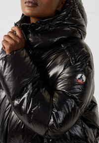 Chaqueta de plumas negra con una textura brillante y suave. Presenta un cuello alto, cremallera visible y un parche con el logo en la manga.