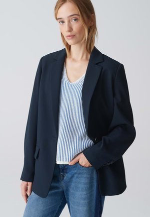 Blazer sartoriale navy con rever a punta, chiusura a un bottone e due tasche frontali, indossato su un top a righe bianche e blu.