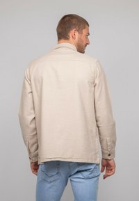 Chemise beige clair à manches longues avec un col, présentant une coupe droite et des poignets boutonnés, associée à un jean bleu clair.
