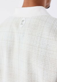 Witte en lichtbeige gestructureerde shirt met een subtiel ruitpatroon, voorzien van een witte kraag en een rond logo-label op de achterkant.