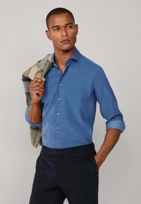 Lichtblauw overhemdd met lange mouwen, één manchet opgerold. Donkere broek. Gekleurd materiaal over de schouder gedrapeerd. Soepele textuur.