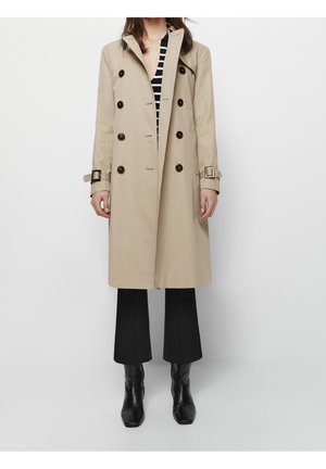 Femme portant un trench-coat beige croisé, pantalon noir court, bottes noires et haut rayé, debout devant un fond uni.