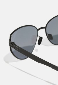 Black sunglasses with dark gray lenses, angular shape, thin metal frame, and textured black temples with a subtle design detail.