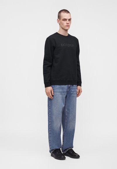 Homme debout portant un sweat-shirt noir avec le texte "DONDUP", un jean bleu ample et des baskets noires sur un fond gris clair uni.