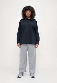 Μαύρη μακρύς μανίκι πόλο μπλούζα με γιακά, γκρι φόρμα και αθλητικά παπούτσια. Η μπλούζα διαθέτει ένα μικρό λογότυπο στο αριστερό στήθος.