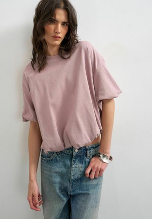 Jeune femme portant un haut ample rose à manches courtes, un jean bleu taille haute et un bracelet manchette argenté, debout contre un mur blanc uni.