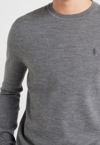 Grå crewneck-tröja i mjukt stickat tyg. Har långa ärmar och en diskret logotypdetalj på vänstra bröstet. Slät textur.