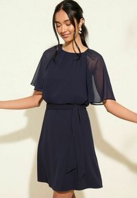Robe bleu marine avec des manches courtes en tulle, un col rond et un détail de taille nouée. Tissu lisse avec un ourlet évasé.