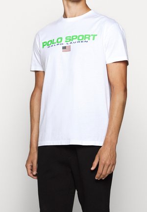 Mężczyzna w białej koszulce na krótki rękaw z nadrukiem "POLO SPORT RALPH LAUREN" i małą amerykańską flagą na piersi.