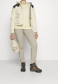 Veste crème claire zippée avec accents noirs, haut beige à manches longues et pantalon marron clair. Bottes de randonnée marron foncé et une casquette claire en main.