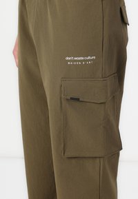 Olive green cargo pants made of soft fabric, featuring a side pocket and white embroidered text "don’t waste culture MAISON D'ART."
