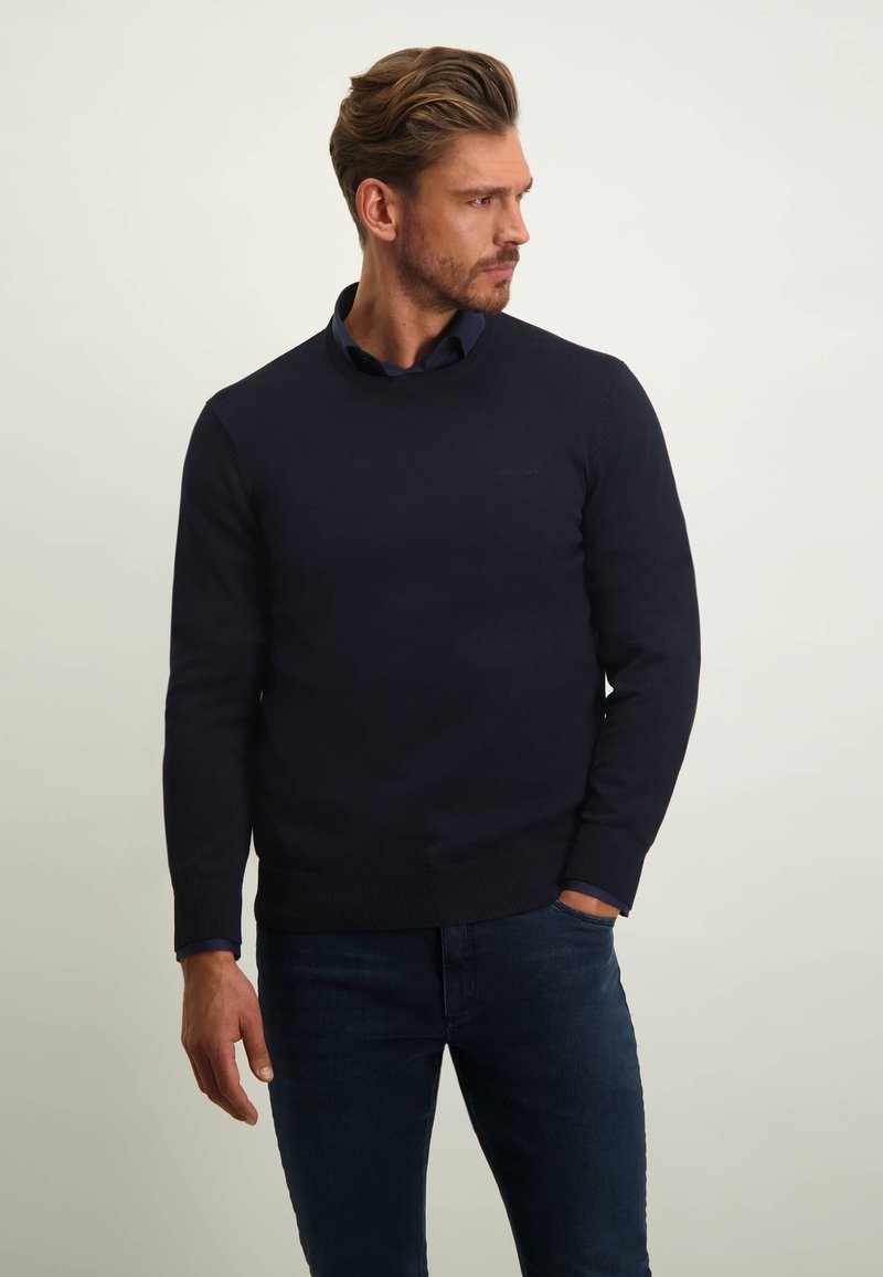 Marineblauer Rundhals-Pullover aus glattem Strick, mit gerippten Bündchen und Saum. Getragen über einem Hemd mit Kragen, kombiniert mit dunklen Jeans.
