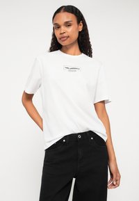 Balta kokvilnas t-kreklā ar melnu uzrakstu "KARL LAGERFELD JEANS". Apvienots ar augsto vidukli melnā džinsos, kuriem ir brīvs piegriezums.