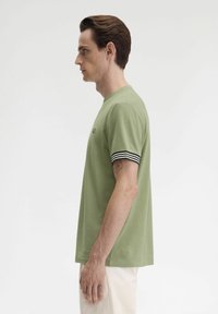 T-shirt verde a maniche corte con polsini a strisce nere e bianche, con colletto rotondo e logo discreto sul petto. Texture uniforme.