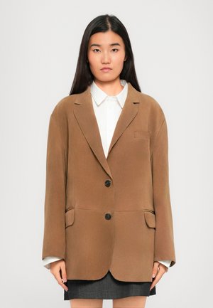 Kvinde med glat, mørkt hår iført en brun oversized blazer over en hvid skjorte med krave og mørk nederdel, stående mod en ensfarvet baggrund.