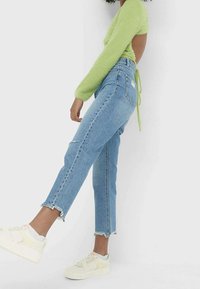 Pull long à manches longues vert clair en peluche, jeans bleus taille haute avec un ourlet effiloché, porté avec des baskets blanc cassé avec des accents pastels.