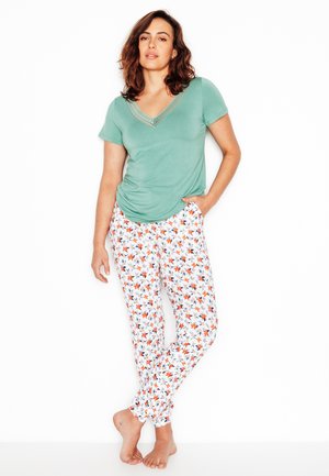 Donna in piedi a piedi nudi con una mano in tasca, indossa una maglietta verde a maniche corte con scollo a V e pantaloni bianchi con motivo floreale.