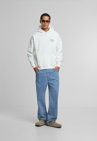 Hvid hættetrøje med en frontlomme og tekstgrafik, parret med lyseblå jeans og tan farvede slip-on sko. Glat tekstur og afslappet pasform.