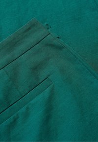 Tkanina w kolorze teal z teksturową powierzchnią, z detalem kieszeni. Materiał wydaje się być miękki i lekko pomięty, z wyraźnymi krawędziami.