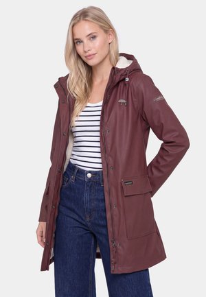 Ung kvinde iført en bordeaux regnfrakke med hætte over en stribet skjorte og blå jeans med den ene hånd i lommen mod en ensfarvet baggrund.