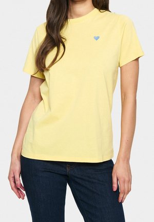 Kvinde iført en ensfarvet gul T-shirt med korte ærmer med en lille blå hjerte-detalje på brystet og mørkeblå jeans.