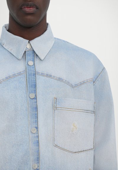 Lys blå denimskjorte med knappelukking foran, lomme med klaff, dobbeltsøm detaljer og subtile slitte detaljer på stoffet og kragen.