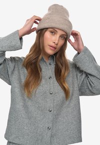 Woman adjusting beige knit beanie, wearing gray button-up sweater with long sleeves, posing against plain light background.