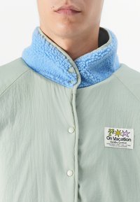 Lichtgroene jas met een blauwe fleecedetail, drukknoopsluiting en een logo patch met kleurrijke iconen aan de linkerkant.