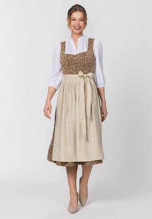 Junge Frau trägt ein braun gemustertes Kleid mit einer weißen Bluse und einer beigen Schürze, die an der Taille mit einer Schleife gebunden ist, lächelnd und stehend.