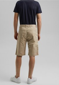 Beżowe szorty wykonane z lekkiego materiału, z bocznymi i tylnymi kieszeniami, o gładkiej fakturze i dopasowanym kroju, zestawione z białymi sportowymi butami.