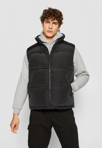 Sort puffer vest med quiltede paneler, lynlås foran og høj krave, lagdelt over en grå hoodie. Stof har en glat, blank overflade.