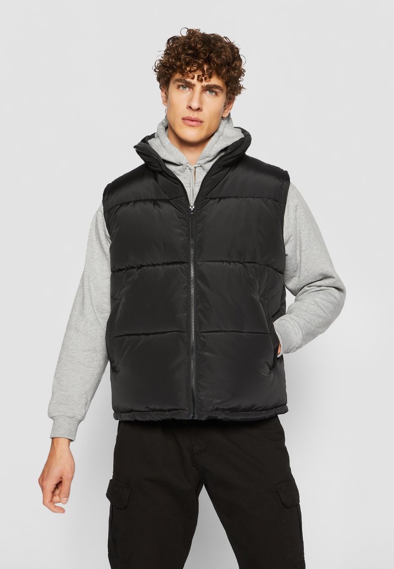 Sort puffer vest med quiltede paneler, lynlås foran og høj krave, lagdelt over en grå hoodie. Stof har en glat, blank overflade.
