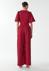 Vrouw met donker haar in een gevlochten bun draagt een op maat gemaakte rode jumpsuit met brede benen en fladderende mouwen, staand tegen een effen witte achtergrond.