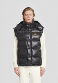 Gilet imbottito nero con design trapuntato, finitura lucida, colletto alto e cappuccio. Presenta tasche frontali e un piccolo patch con logo sul petto.