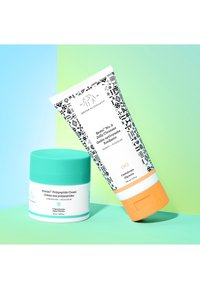 Two skincare products on a gradient background. A teal-topped jar with white label and a patterned white tube with orange base and cap.