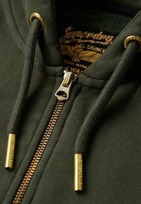 Sudadera verde oscuro con cremallera hecha de material suave. Presenta una cremallera metálica con acentos dorados y puntas de cordón doradas. Logo de la marca visible en el interior.