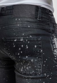 Černé džíny z denimu s vybledlým a opotřebovaným vzorem s bílými skvrnami od barvy. Obsahují koženou nášivku na opasku a zadních kapsách.
